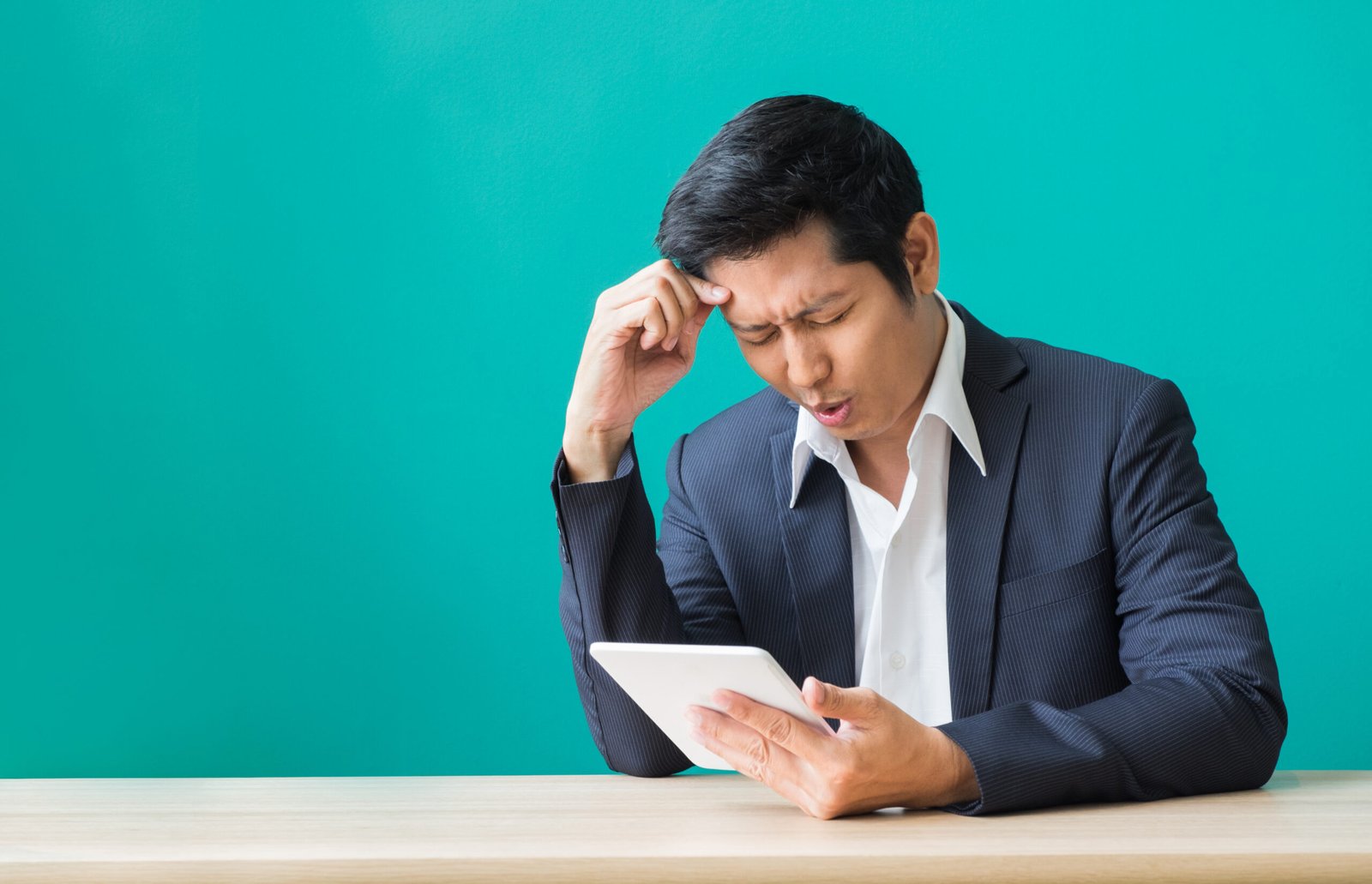 Homme en costume fronçant les sourcils et se grattant la tête en regardant une tablette, exprimant la confusion et l’incertitude.