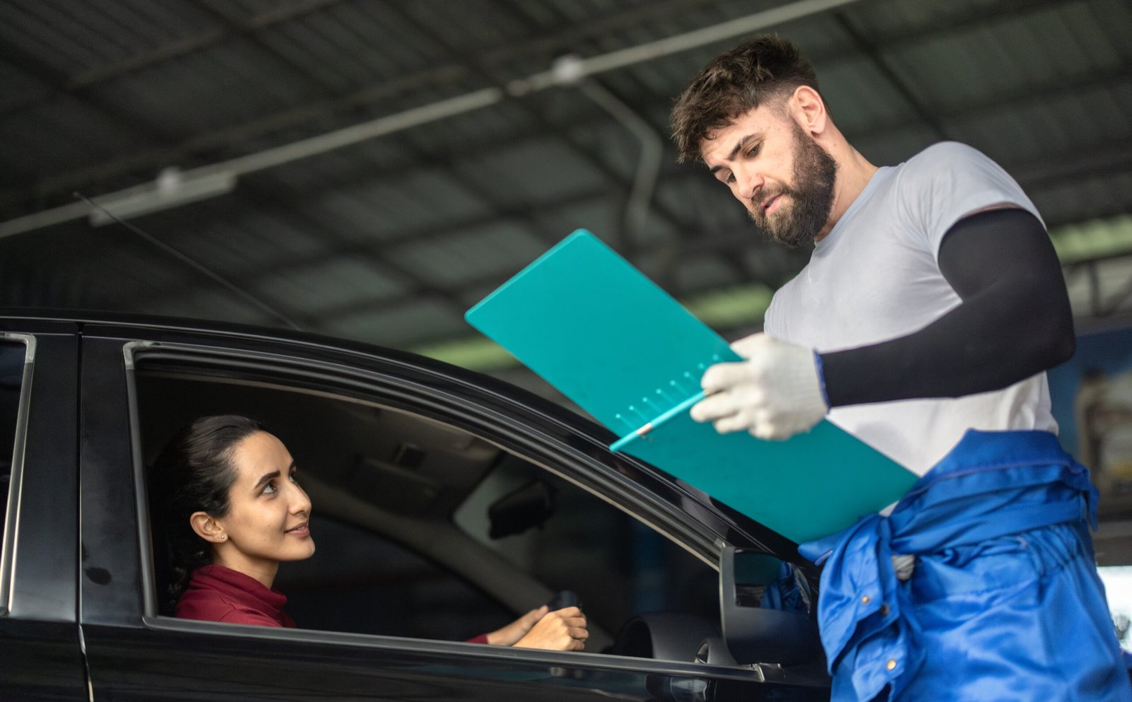 Garagiste en tenue bleue présentant un document à une conductrice assise dans sa voiture noire.