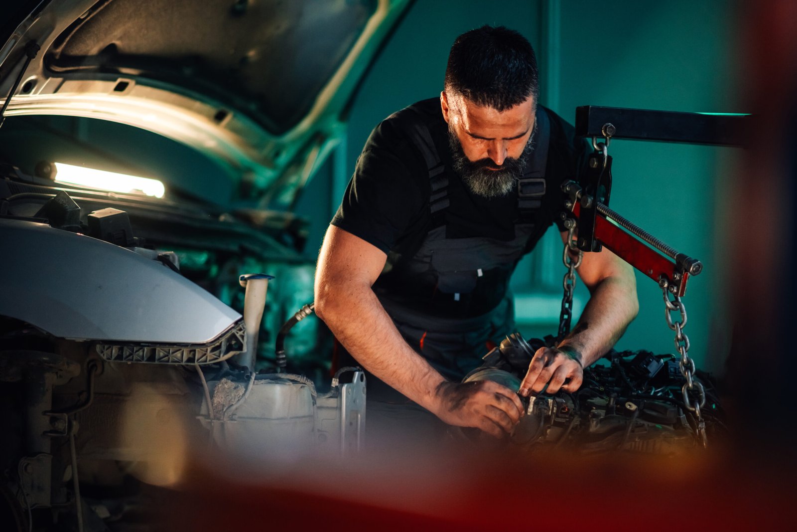 Mécanicien barbu en combinaison noire réparant un moteur démonté dans un garage.