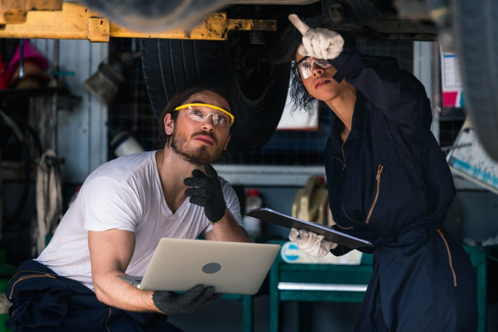 Deux mécaniciens effectuent un diagnostic automobile sous un véhicule, l’un tenant un ordinateur portable et l’autre pointant un problème tout en tenant un rapport.