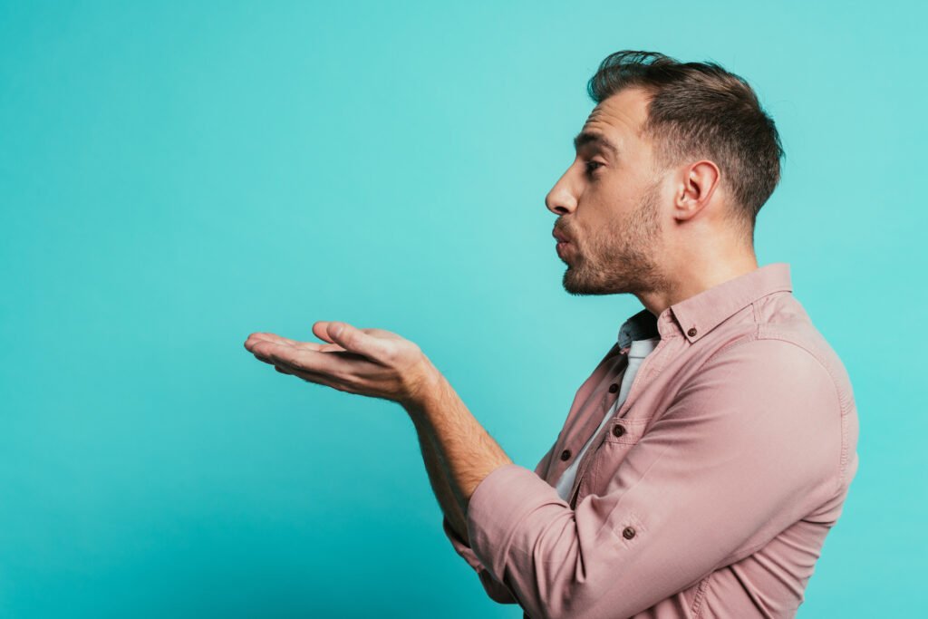 Homme en chemise rose faisant un geste de souffle symbolique avec ses mains, sur fond turquoise.