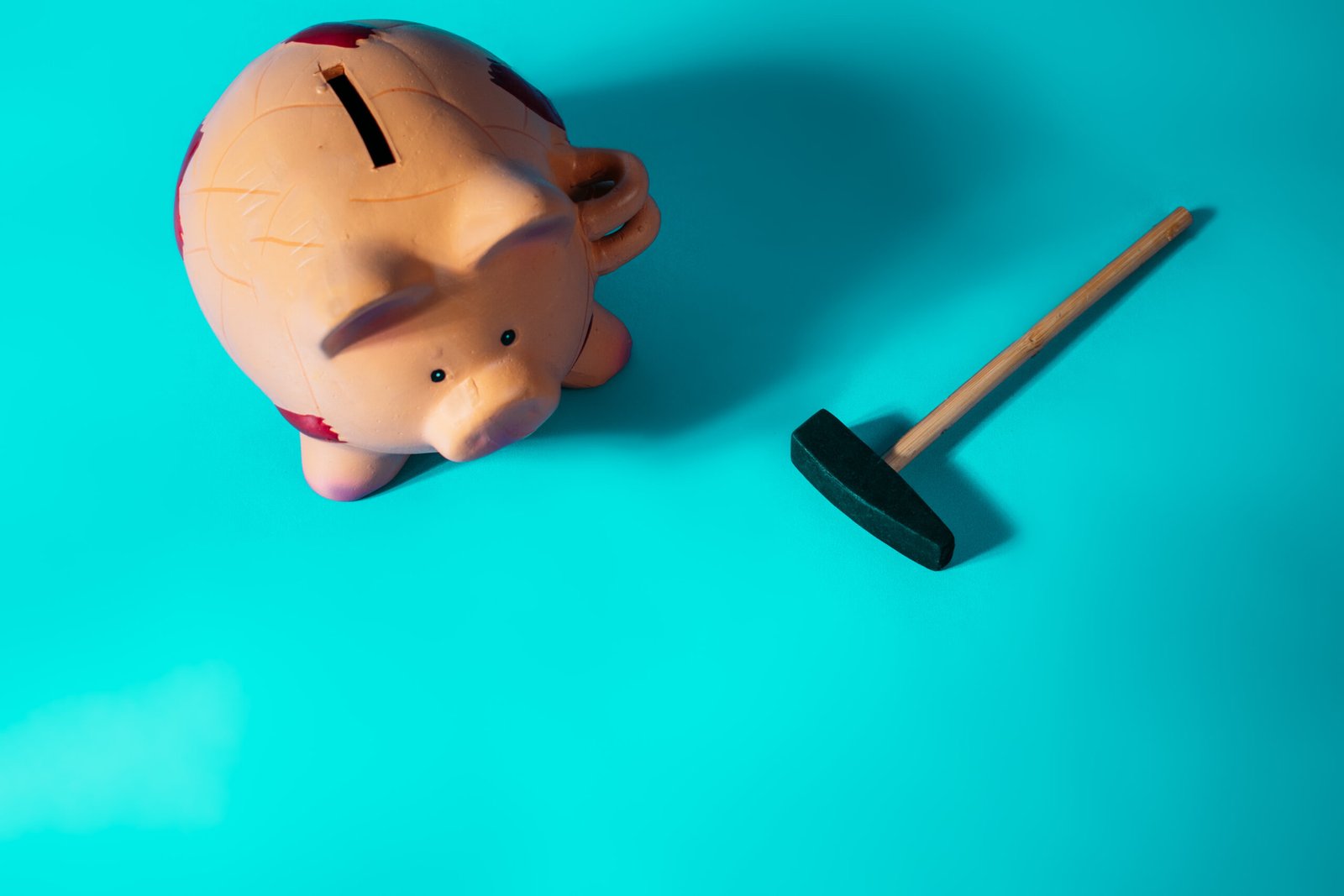 Tirelire cochon posée sur une surface turquoise avec un marteau à côté, symbolisant la pression financière et les économies menacées.
