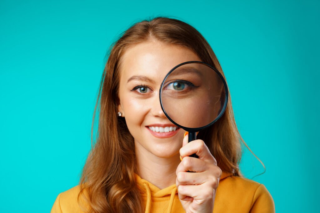 Femme souriante tenant une loupe devant son œil, symbolisant l’analyse minutieuse et la recherche de détails lors d’un diagnostic ou d’un litige automobile.
